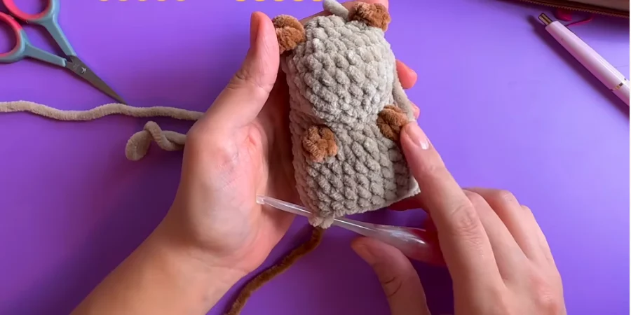 Hands holding a partially completed gray crochet capybara with brown ears and legs, while a crochet hook is used to work the lower edge on a purple surface.