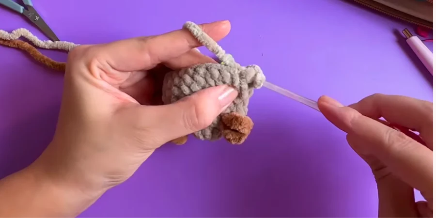 Hands holding a partially completed gray crochet capybara while adding a brown crocheted detail with a crochet hook.