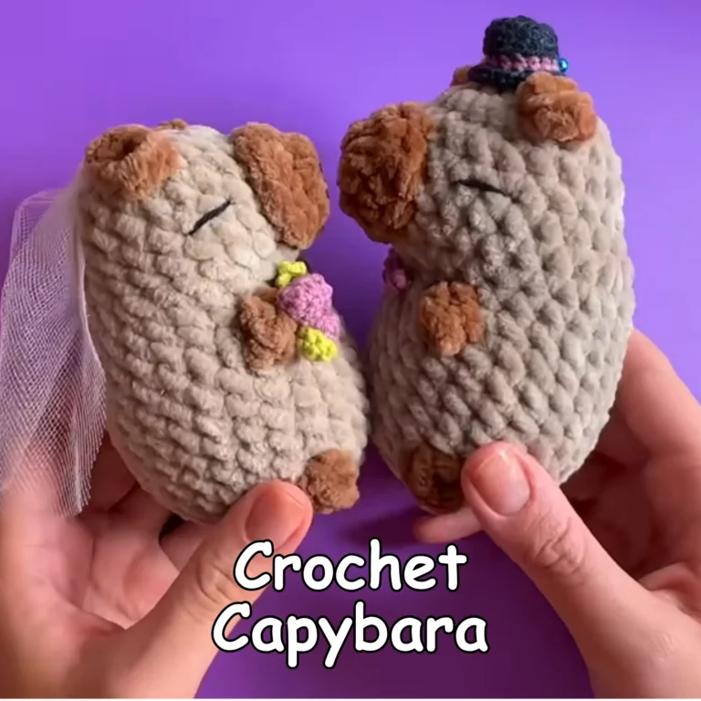 Two finished crochet capybara plushies displayed together, featuring gray bodies, brown ears and feet, with one wearing a small black hat and the other decorated with a pink flower.