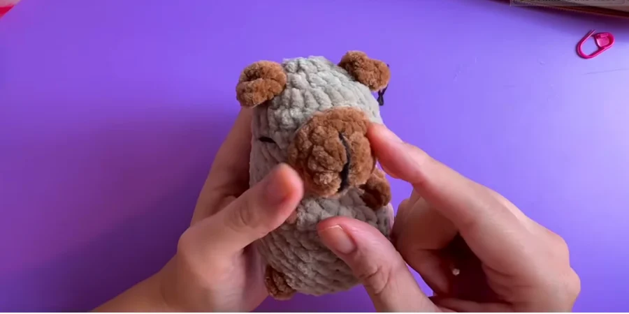 Close-up of a finished gray crochet capybara plush with brown ears, snout, and feet, held in both hands against a purple background.