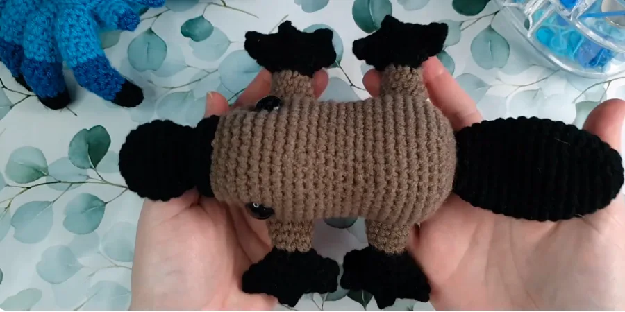 Completed crocheted platypus showing the brown body, black bill, tail, and four black feet during assembly.