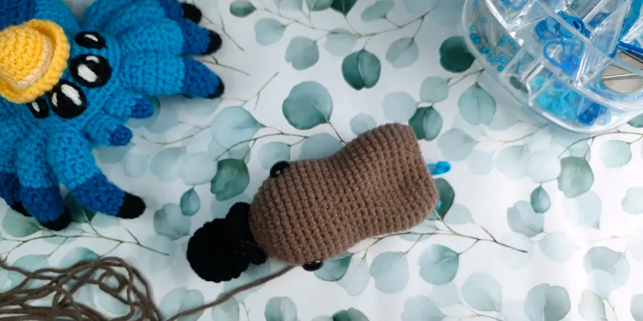 Brown crocheted platypus body shown from the side with safety eyes and black tail attached.