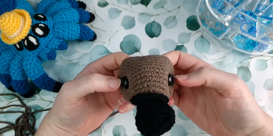 Close-up of a crocheted brown platypus head with black safety eyes and the black bill being attached.