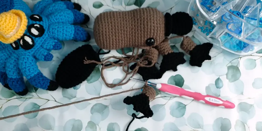 Platypus crochet pattern in progress with brown body, black feet and tail pieces, crochet hook, yarn, and stitch markers on a work surface.