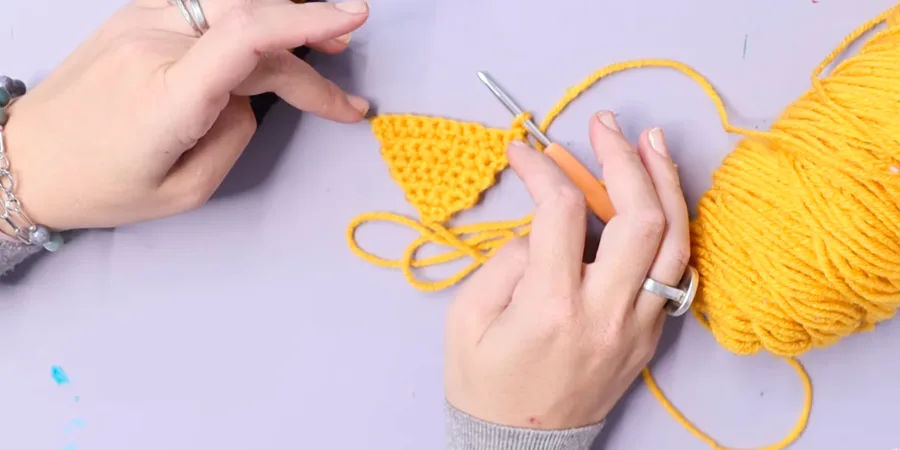 A yellow crocheted dreidel middle piece shown in progress with a crochet hook and working yarn.