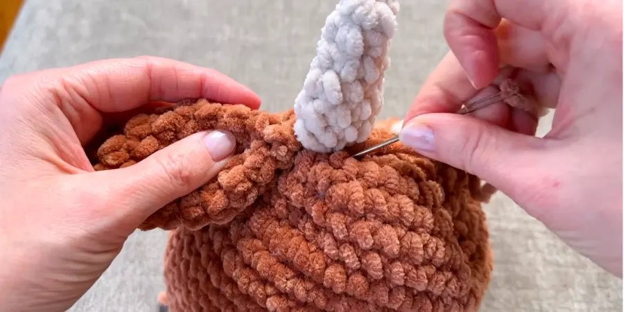 Attaching a white crocheted ear to the brown Highland cow body with a yarn needle.