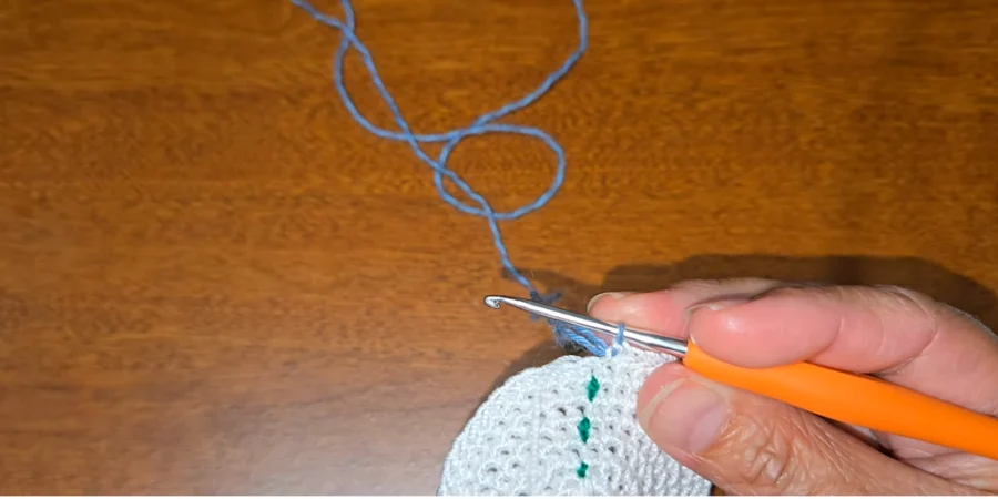 Close-up of a crochet hook working the edge of a white circular kippah with a green yarn marker.
