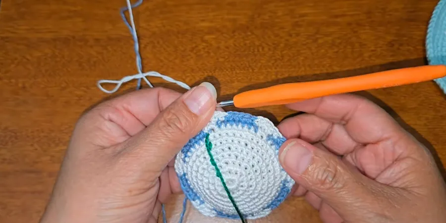 Hands holding a partially crocheted white kippah with blue colorwork around the outer edge and a green round marker.