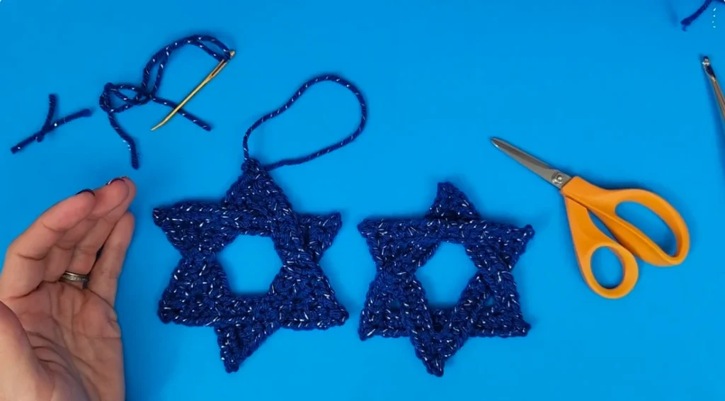 Two completed blue crocheted Star of David ornaments displayed side by side, with loose yarn tails, a yarn needle, and scissors nearby.