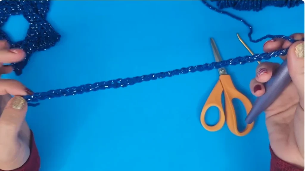 A long blue crochet foundation chain being held beside a crochet hook and scissors during the beginning of the pattern.