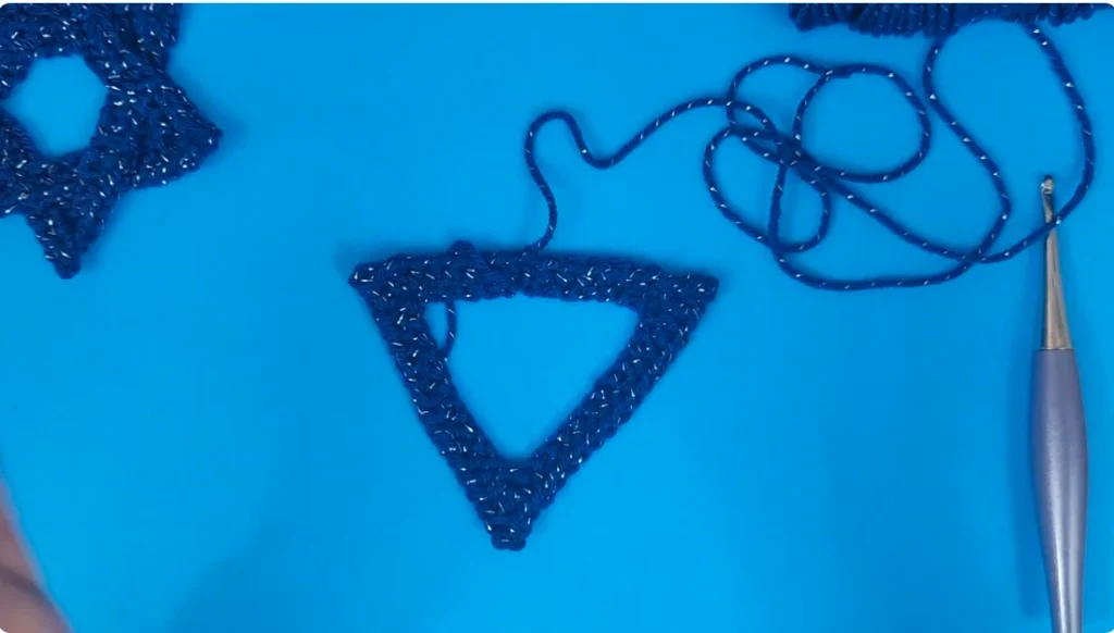 A single blue crocheted triangle completed with three pointed corners and an open center, with a crochet hook and yarn beside it.