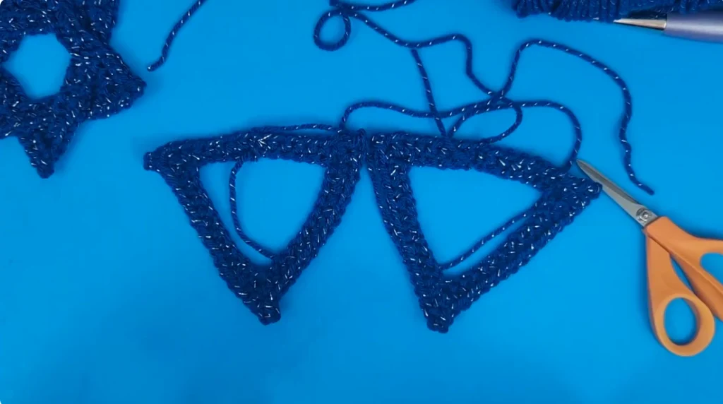 Two blue crocheted triangles positioned side by side before being interlocked to form a Star of David, with scissors and yarn visible