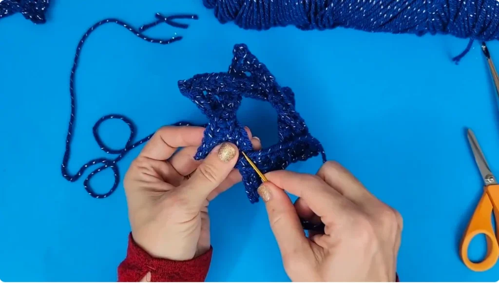 Hands securing the overlapping sections of a blue crocheted Star of David using a yarn needle, with scissors and yarn nearby