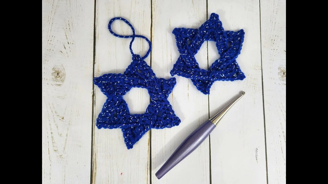 Two finished blue crocheted Star of David ornaments with silver metallic accents, displayed on a white wooden surface beside a crochet hook.