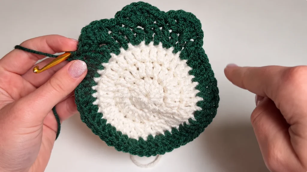 Close-up of a crocheted cream-and-green circular motif with a gold crochet hook and hands demonstrating the edging