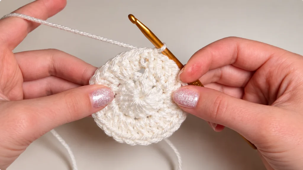 Close-up of hands working a small cream crochet circle with a gold hook and white yarn.
