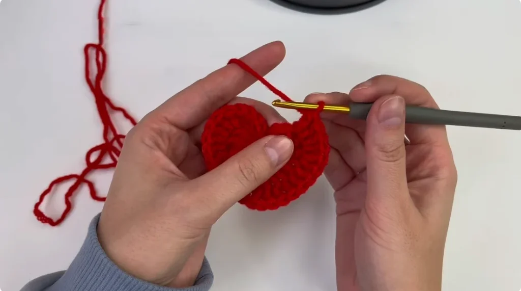 Hands crocheting a red heart motif with a gold crochet hook and red yarn