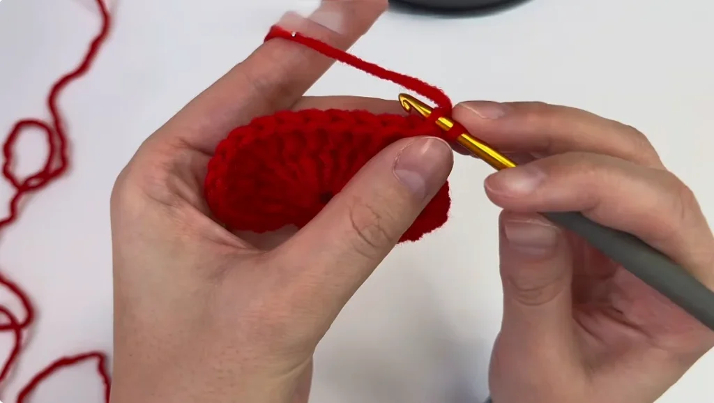 - Close-up of hands using a crochet hook to work red yarn along the edge of a heart motif.