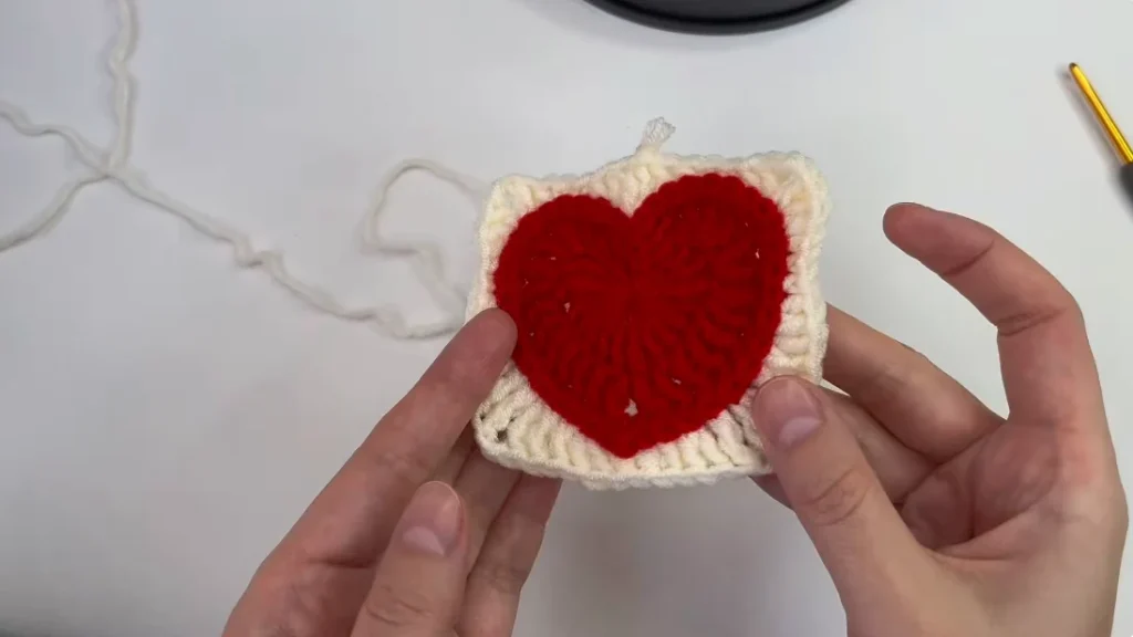 Red crocheted heart granny square with a cream border, held in both hands during the finishing stage.