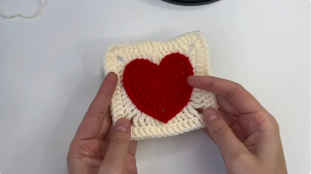 Finished red heart granny square with a cream crochet border, held up to show the completed design.