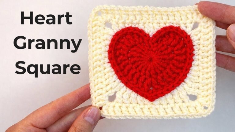 A red crocheted heart granny square with a cream-colored border, featuring a textured heart centered within the square. The motif is held up by hand against a light background.