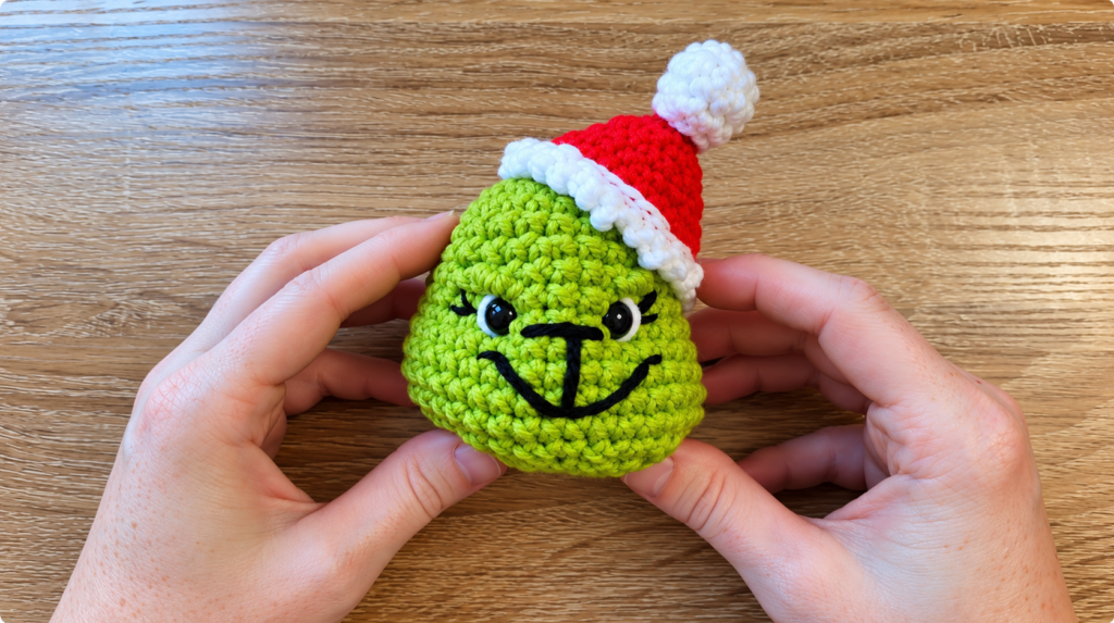 Finished Grinch crochet ornament with Santa hat — Hands holding a green crocheted Grinch ornament wearing a red-and-white Santa hat.