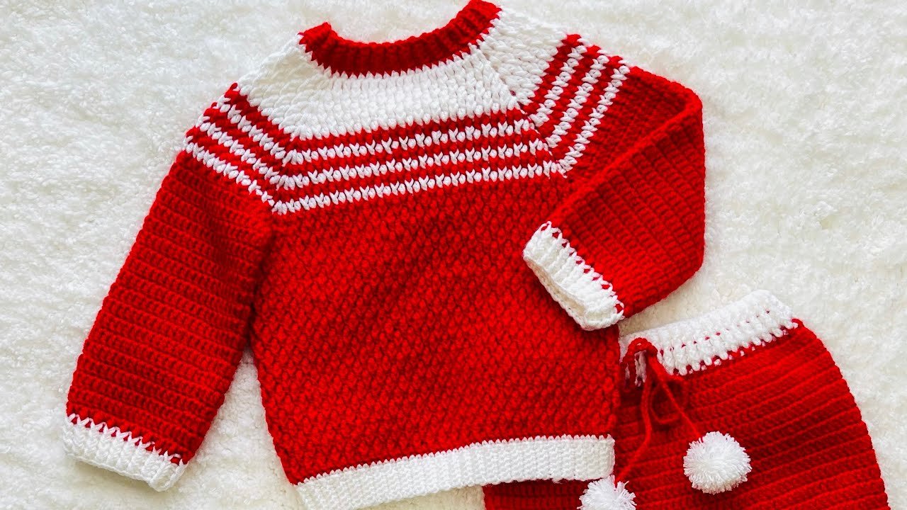 A red and white crocheted Christmas baby sweater with a textured yoke, red ribbed sleeves, white cuffs and neckline, displayed on a soft white background.
