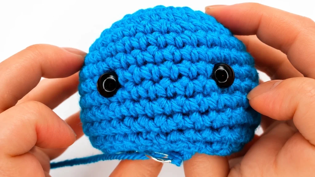 Close-up of a blue crocheted snowman head with two black safety eyes being positioned by hand.